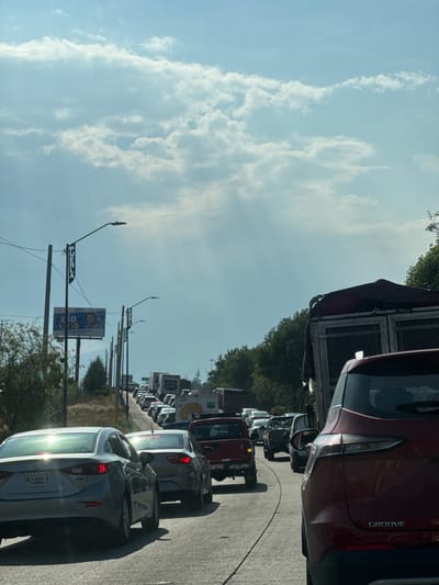 The image shows a long line of stopped or slow-moving traffic on a road in San Pedro Cholula, Mexico. Cars and trucks of various sizes and colors are visible. The sky is partly cloudy. A sign reading "Puebla" can be seen attached to a street lamp post. A billboard is also visible in the background. The license plate on the closest car reads "TWJ-664-A". A vehicle with the name "GROOVE" printed on its side is in the upper right part of the frame. The environment includes trees and vegetation lining the roadside.