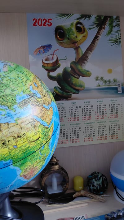 The image displays a corner of a room with various objects on a shelf. A large globe, showing parts of Europe, Africa, and the Middle East, is prominently placed on the left. To its right, a calendar for the year 2025 is visible, featuring a cartoon illustration of a snake on a palm tree, holding a coconut drink with a small umbrella. The calendar has sections with dates in Armenian and Russian. On the shelf below, there's a decorative lantern, a yellow object, a dark, textured sphere, a feather, and some thin wooden sticks. A portion of a blue and white lamp is seen on the far right. The background appears to be a light-colored wall or shelf. There are no people in the image. The presence of Armenian text on the calendar strongly suggests a connection to Armenia, and specifically to the city of Abovyan, given the context provided. The arrangement of objects suggests a personal collection or display.