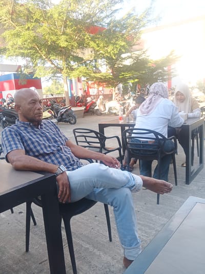 The image depicts an outdoor scene at what appears to be a roadside cafe or rest stop in Geulumpangsulu Timu, Indonesia, with a gas station in the background. In the foreground, a bald, middle-aged man with dark skin is seated at a table, looking to his left. He is wearing a blue and white checkered short-sleeved shirt and light blue jeans. His left leg is crossed over his right, and he is wearing a silver watch on his left wrist.
Behind the man, at another table, two women are seated. One woman, wearing a light-colored hijab and a white long-sleeved shirt, is looking down at the table. The other woman, also wearing a hijab and a tan long-sleeved top, is seated next to her, also looking towards the table. A child is partially visible between them. In the background, several motorcycles are parked, and the structure of a gas station is visible with red and blue accents. The scene is lit by bright sunlight, suggesting it is daytime.