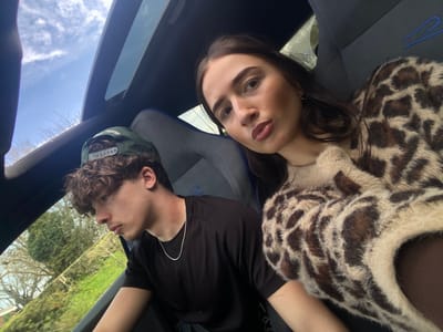 The image displays two individuals seated inside a vehicle. On the right, a female with long dark hair and light complexion is visible from the chest up, wearing a light-colored, fluffy sweater with a dark brown leopard print pattern over a dark garment. She gazes directly towards the camera with slightly pursed lips. On the left, a male with curly brown hair and a light complexion is present, wearing a dark green baseball cap reversed, a plain black t-shirt, and a thin silver chain necklace. He is positioned looking downwards and to his left, away from the camera.
Both individuals occupy what appear to be the front seats of a vehicle, which features dark upholstery with blue accent stitching visible on the seatbacks. An open panoramic sunroof above reveals a clear blue sky with scattered white clouds. Through the vehicle's windows, a backdrop of green foliage, including trees and bushes, is visible, indicating an outdoor setting. While the prompt specifies Broxbourne, United Kingdom, no discernible landmarks or unique features are present within the image to forensically confirm this precise geographical location.