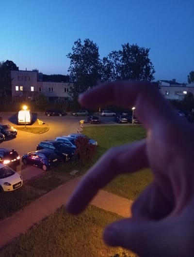 The image depicts an outdoor scene at dusk or early evening in Radzymin, Poland. The sky is a deep blue, indicating the transition from day to night. In the foreground, a blurry hand is positioned in a way that it partially obscures the view of the scene below.
The scene itself features a residential area with multi-story buildings visible in the background. Some windows in the buildings are illuminated, suggesting people are inside their homes. A street or parking area runs horizontally across the mid-ground, lined with parked cars of various colors, including dark blue, black, and white. A lamppost illuminates a section of the parking area. Adjacent to the parking area is a grassy lawn. A pathway, possibly made of pavers, runs along the grass and parking area, illuminated by the ambient light. Trees are also present, with one large tree on the left side of the frame and smaller trees or bushes near the buildings and along the pathway.
The overall impression is a quiet, residential evening. There are no discernible emotions conveyed due to the blurriness and focus on the environment. No text is visible in the image. The presence of parked cars and residential buildings suggests a typical urban or suburban setting.