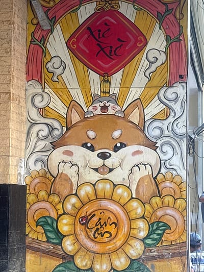 The image displays a vibrant, cartoon-style mural painted on an exterior wall in Thuận Hóa Ward, Vietnam. The mural's central figures are a large, plump, brown and white dog, resembling a Shiba Inu, with a wide, happy smile showing its tongue and raised front paws. Perched on the dog's head is a smaller, grey and white cat, also depicted with a wide, open-mouthed smile. Above them, a red diamond-shaped banner, featuring black Chinese characters "謝謝" (xièxiè), commonly used for "thank you" in Vietnamese contexts, hangs from a golden tassel. This banner is set against a yellow and white sunburst background, flanked by decorative red scrollwork and swirling grey clouds. The bottom portion of the mural showcases a row of bright yellow sunflowers with green leaves. The largest central sunflower features the Vietnamese text "Cảm ơn" (Thank you) written in black script within its center, accompanied by a small red ladybug on one petal. To the left of the mural, a vertical column of light brown, textured bricks is visible, exhibiting some surface wear. To the right, multiple black electrical wires are bundled and extend vertically against a light-colored background, possibly a metal roll-down door. A person wearing a blue baseball cap and a striped shirt is partially visible in the lower right corner, facing away from the viewer. The mural itself shows some localized dirt and minor discoloration.