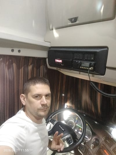 A man with short dark hair and a light complexion is pictured from the chest up, looking directly at the camera. He is wearing a white t-shirt and is seated in the driver's seat of a truck. His right hand is raised and positioned near the steering wheel, with two fingers extended in a peace sign. The steering wheel is black with silver accents and the "DAF" logo is visible. The dashboard of the truck is also visible, featuring various gauges and buttons. Above the dashboard, a CB radio is mounted. The background consists of brown and black striped curtains and the interior ceiling of the truck cab. A timestamp in the lower left corner reads "2026.06.21 11:55". The image appears to have been taken inside the cab of a DAF truck, likely on a road or at a truck stop. There is no information to indicate this location is in Gorizia, Italy.