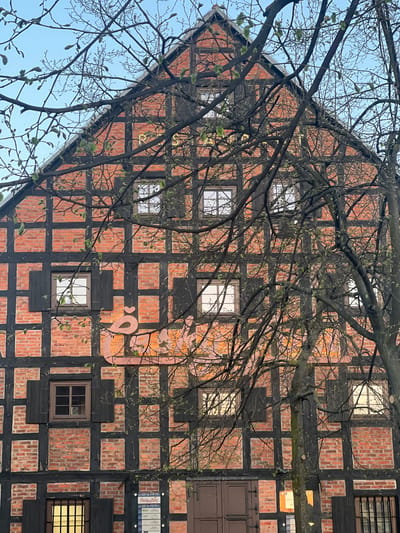 The image depicts a fragment of a brick building with dark timber framing, partially obscured by tree branches. The building has several windows, some of which are lit from within. A decorative element, possibly a painted sign or symbol, is visible on the brick facade. The architecture suggests a historical building. The photo is taken from an unusual angle, looking upwards. Based on the architectural style and color of the brick building, the image is likely located in Bydgoszcz, Poland. The photograph depicts the facade of the Leon Wyczółkowski District Museum, Bydgoszcz, Poland.
