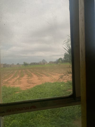 The image captures a view through a screened window of a rural landscape in Tavo, Nigeria. The foreground is dominated by the mesh of the window screen, which casts a subtle grid over the scene. Beyond the screen, a field with rows of crops is visible, interspersed with patches of orange soil. In the distance, a cluster of buildings, possibly houses, can be seen, along with some trees. The sky is overcast, suggesting a cloudy or potentially rainy day. The overall impression is one of a quiet, agricultural setting.