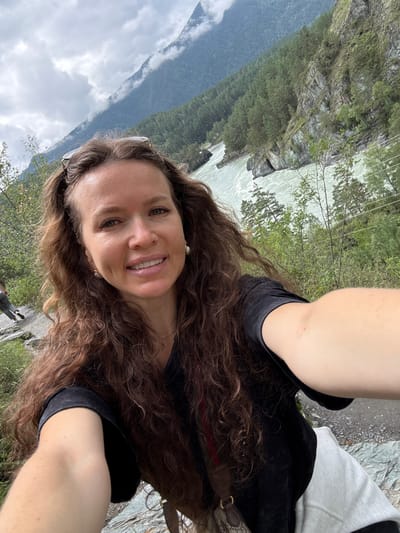 In a selfie taken outdoors, a woman with long, wavy brown hair smiles at the camera. She is wearing a black t-shirt and a light-colored hooded garment. Sunglasses are perched on top of her head. Behind her, a wide, turquoise river flows swiftly through a rocky gorge. Steep, tree-covered slopes rise on either side of the river, and dramatic, cloud-shrouded mountains loom in the background. A second person, whose legs and lower body are visible, is walking away from the camera on a path to the left. The scene appears to be in the mountainous region near Chemal, Russia, likely along the Katun River, a popular tourist destination known for its scenic beauty.