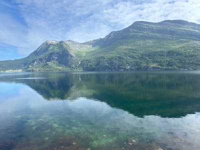A reflective, wide shot captures a serene Norwegian fjord landscape under a partially cloudy sky. The scene is dominated by a vast, calm body of water in the foreground, its surface mirroring the lush green mountains and blue sky above. The water is clear enough in the lower part of the frame to reveal the rocky, weedy bottom. Beyond the water, imposing, verdant mountains rise, their slopes densely covered in trees. A thin waterfall cascades down a rocky incline on the left side of the mountains. On the far left, at the base of the mountains and near the water's edge, a small cluster of buildings, likely houses, is visible, suggesting the presence of a settlement in the city of Inndyr, Norway. The overall impression is one of natural beauty and tranquility.