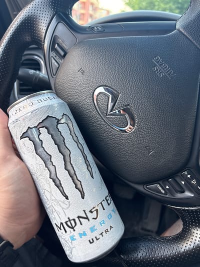 An eye-level shot inside a car, focusing on a hand holding a Monster Ultra energy drink can, with a steering wheel dominating the background. The scene appears to be set in Plovdiv, Bulgaria, under bright daylight conditions.
The main subjects are a person's hand holding a white and silver Monster Ultra energy drink can, and a black leather steering wheel. The steering wheel, likely belonging to an Infiniti vehicle as suggested by the prominent chrome logo in the center, features various control buttons. These include audio controls with "+" and "-" for volume and a "SOURCE" button, as well as cruise control settings labeled "COAST/SET", "ACCEL/RES", and "CANCEL". Additional buttons for "ON" and "OFF" are visible, possibly related to cruise control or other functions. The text "SRS AIRBAG" is embossed on the steering wheel's central pad, indicating the presence of an airbag. Parts of the steering wheel have a perforated leather texture.
The Monster Energy can is clearly visible, showcasing its distinctive claw-mark logo in silver against a white background with subtle patterns. Text on the can reads "MONSTER ENERGY ULTRA" and "ZERO SUGAR". The activity depicted is simply holding the energy drink within the car environment, possibly while driving or parked. No other people are visible, and emotions cannot be discerned.