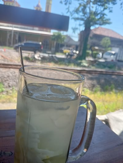 A tall, clear glass of iced beverage, possibly a local Indonesian drink like kelapa muda (young coconut), sits on a wooden surface. The glass contains ice cubes and what appears to be shredded coconut flesh, with a silvery straw or stirrer inside. The background is blurred but suggests an outdoor setting. Visible elements include train tracks, a dark-colored car parked near a building with arched windows, other vehicles, trees, and buildings with tiled roofs. The sky is clear and blue. The overall impression is a casual scene, likely taken at a roadside establishment or a cafe near a railway line in the area of Sruni, Indonesia.