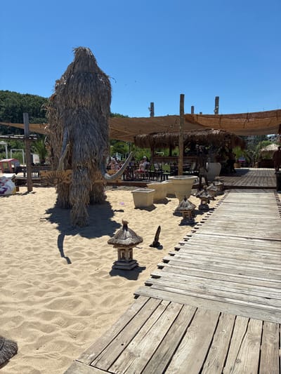 The image depicts a sunny outdoor scene on a sandy beach, likely at a beachfront establishment in Shkorpilovtsi, Bulgaria. In the foreground, a large, rough-textured mammoth-like sculpture made of dried grass and straw stands prominently on the sand. It has two visible tusks and a wooden sign with an eye and sun emblem attached to its body. To the right of the sculpture, a weathered wooden boardwalk extends diagonally into the scene, with several small, pagoda-style decorative structures placed along its edge.
In the midground, a shaded area with thatched roofing and wooden support poles is visible, housing tables and chairs, with several patrons seated and enjoying the atmosphere. Beyond this, more of the beach and vegetation can be seen. The sky is a clear, bright blue. The overall impression is that of a relaxed, resort-like environment.