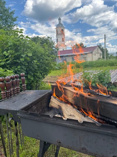 The image captures an outdoor scene in what appears to be a village setting, likely in Луптюг, Russia, given the presence of a distinct church tower. In the foreground, a metal barbecue grill is set up, with logs burning intensely, producing bright orange flames and wisps of smoke. Several wooden skewers with carved handles are neatly arranged on the side of the grill. The grill is placed on a patch of green grass. In the middle ground, a simple building with a red roof is visible, and further in the background, the aged, weathered stone tower of a church stands tall against a bright blue sky dotted with fluffy white clouds. Lush green foliage from trees and bushes surrounds the area, suggesting a rural or semi-rural environment. The overall impression is one of a peaceful, perhaps summery, day. There are no people visible in the image.