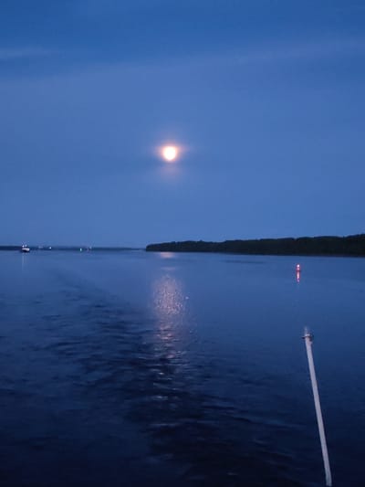 The image depicts a body of water, likely a river or sea, at twilight. A bright, full moon is visible in the deep blue sky, casting a soft glow. In the distance, a dark line of trees forms the shoreline. The water's surface shows ripples and a reflection of the moon. A white pole extends vertically from the bottom right corner of the frame, suggesting it is attached to a boat or dock. Several small lights are scattered across the water in the distance, possibly from other vessels or navigational markers. The scene is serene and tranquil. The location is described as being in Matveevskoe, Russia.
