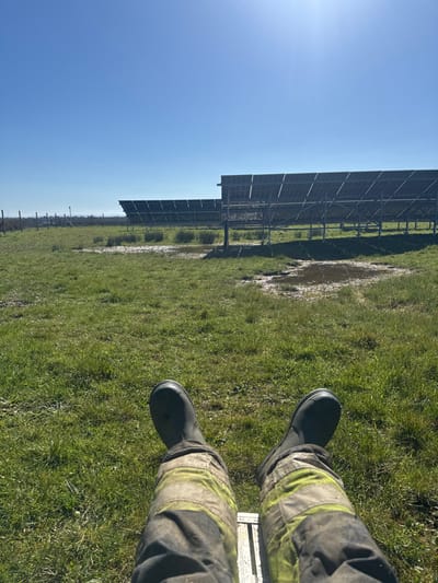 A first-person perspective image depicts the lower extremities of an individual within the Millbrook Solar Farm in Southampton, United Kingdom. The foreground shows soiled dark grey work trousers featuring prominent yellow-green reflective bands, paired with black rubber boots. The individual's legs are extended, suggesting a seated or reclined position, possibly on a metallic surface visible beneath the calves. The midground and background consist of a large, open grassy field under a clear blue sky. Numerous muddy puddles are distributed across the grass. Multiple rows of industrial-scale solar panels are erected across the field, viewed from their rear support structures. The sun, located in the upper right, produces a noticeable flare, and a faint aircraft contrail is visible in the upper left sky.