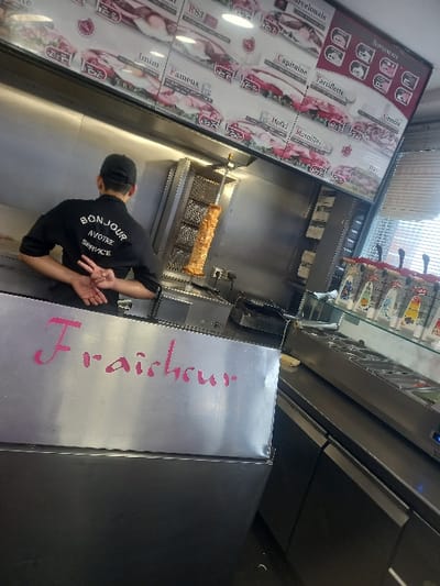 An interior view of a fast-food establishment, located in Sallaumines, France, features a male employee seen from behind, wearing a black uniform shirt with "BONJOUR À VOTRE SERVICE" printed on the back and a black baseball cap. His hands are clasped behind his back, with the left hand displaying a peace sign. Directly in front of the employee is a vertical rotisserie holding a large, cooked doner kebab skewer. Above this, a prominent illuminated menu board displays various food items, depicted with images and text in French, alongside prices. The background wall behind the rotisserie is clad in polished stainless steel panels. In the immediate foreground, a stainless steel counter features the word "Fraîcheur" (Freshness) written in pink, stylized script. To the right of the scene, an extended stainless steel counter holds embedded food preparation pans and a tiered display of clear plastic squeeze bottles containing sauces. A window with horizontal blinds is visible on the far right, admitting external light. The environment is functional, characterized by commercial kitchen equipment and stainless steel surfaces.