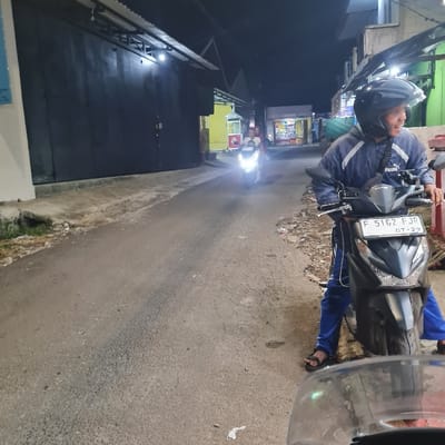 The image shows a man sitting on a black motorcycle in a narrow street in Kecamatan Tamansari, Indonesia (latitude: -6.638, longitude: 106.735). He is wearing a blue jacket with a white Puma logo. He is wearing a black helmet with a white visor and blue jeans. The motorcycle has a black license plate with the number "F 5162 FJP" and "07-29" printed below it.  The man is looking directly at the camera.  The motorcycle is facing the camera. The man is sitting on the motorcycle, his right leg is on the ground, and his left leg is on the motorcycle's footrest.  There is another motorcycle in the background, heading toward the camera.  The street is asphalt and it is dark. There are buildings on both sides of the street.  The buildings are made of concrete and have white paint.  The street is empty except for the man on the motorcycle.