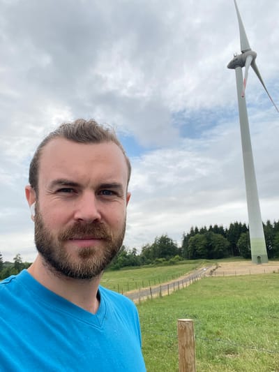A man with blonde hair pulled back and a beard is taking a selfie in front of a wind turbine in Schwarzenborn, Germany. He is wearing a blue t-shirt and white earbuds. The wind turbine is tall and white with a green base. In the background, there is a field, a dirt road, a line of trees, and a cloudy sky. The man is the main subject of the image.
