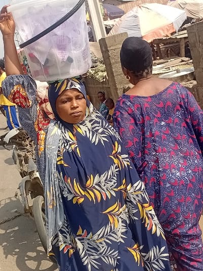 Two women are outdoors, one in a patterned blue and yellow hijab-like head covering, with a clear plastic container holding wrapped items next to her. She faces the camera, wearing colorful, patterned clothing. A second woman stands behind her, wearing a red and dark blue patterned dress. The background shows a building under construction and general outdoor clutter, along with another person partially visible further back.