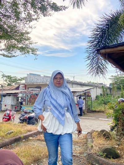 An adult female stands facing the camera in an outdoor setting in Kalitengah, Indonesia. She wears a light blue patterned hijab, eyeglasses, a light blue long-sleeved top featuring a white puffy lower section with small white bows, and light blue jeans. Her right hand holds a dark, slender object.

The background includes various elements:
To her immediate left, two motorbikes are parked, a red Honda Scoopy scooter and a black motorbike. Behind them, multiple informal structures with corrugated metal roofs are visible, consistent with market stalls or small shops. A male individual in a blue collared shirt stands with his back to the camera among these structures. Further back, a gray concrete wall or bridge spans horizontally across the scene, with power lines overhead.

To her right, a rustic wooden beam structure with a portion of a roof is visible, along with green foliage and palm fronds in the upper right. The ground in the foreground consists of dry earth and sparse grass, delineated by wooden logs or beams. The sky above is partly cloudy blue.