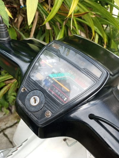 The image displays a close-up view of the dashboard of a black and white scooter or moped, situated in Salai Har Htan, Myanmar. The main subject is the instrument cluster, featuring a speedometer and fuel gauge under a clear, somewhat scuffed plastic cover. The speedometer needle rests at zero, and the odometer displays "00000" in red. Indicators for "FUEL," "OIL," and "HI BEAM" are visible. Below the instrument cluster, an ignition switch is present, labeled "OFF" and "ON," with "IGNITION" printed at the bottom; no key is in the keyhole, and the switch appears to be in the "OFF" position. Two corroded screws secure parts of the dashboard. The scooter's top fairing is black plastic, transitioning to white bodywork in the lower foreground. A black cable is visible entering a hole on the right side of the dashboard. The background consists of out-of-focus green and yellow-green foliage, suggesting an outdoor environment, with a light-colored ground surface partially visible on the bottom left. No individuals or active interactions are depicted.
