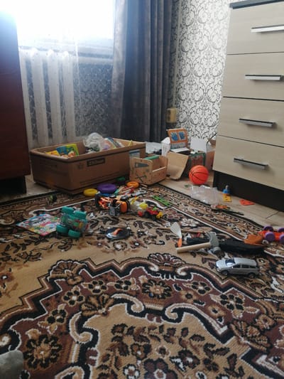 The image shows a child's bedroom in Dubrovka, Russia, with toys scattered across a patterned rug. The scene is cluttered, suggesting recent play. A large cardboard box, labeled "250мл" and with Russian text, is filled with various toys, including soft blocks and plastic items. Other toys are spread out on the floor, such as a green toy truck, a toy car, a basketball, and a toy hammer. In the background, a window with sheer curtains is visible, letting in natural light, and a tall dresser with wooden drawers stands against a wall decorated with a grey and white floral wallpaper. The overall impression is one of a lived-in space, typical of a child's play area. The time of day appears to be daytime, as suggested by the natural light. There are no people visible in the image.