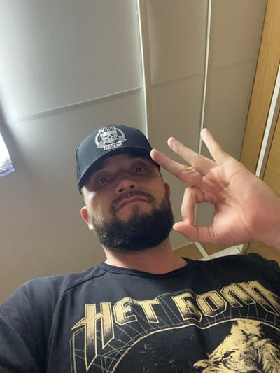A man with a beard is angled in the frame, wearing a black t-shirt with a gold design and a black baseball cap with a white logo. His right hand is raised, fingers spread. The background consists of a light-colored wall with horizontal lines. The location information (city of Koskolovo, Russia) is not discernible from the image.