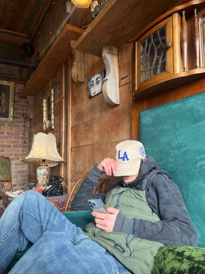 A person is seated in a deep green upholstered armchair within an establishment in Argveta, Georgia. The individual wears a beige baseball cap with "LA" in blue lettering, a dark grey long-sleeved shirt, a green quilted vest, and blue jeans. The cap is pulled low, obscuring the face, as the person holds and looks at a light-colored smartphone with both hands.
The room features rustic wooden wall paneling and a section of exposed red brickwork on the left. A wooden shelf supported by two carved corbels runs across the upper wall. Decorations include a small framed painting of a face with prominent eyes positioned between the corbels, a larger framed classical painting on the brick wall, and a small rectangular mirror. To the left of the seated person, a vintage table lamp with a lace-patterned shade and a floral base sits on a small table or stand, alongside a black rotary telephone and an indistinct dark object, all resting on a patterned rug. In the upper right corner, an antique-style wooden cabinet with a leaded glass door and carved vertical elements is visible. The ceiling incorporates exposed wooden beams and an ornate light fixture.