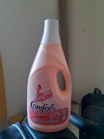 A bright pink plastic bottle of Comfort fabric conditioner stands upright on a dark surface, possibly a shelf or furniture. The bottle has a white cap and a large, oval-shaped handle integrated into its side. A label on the bottle features a graphic of a woman in a flowing pink dress with her arms outstretched, seemingly dancing. The text "Comfort" is prominently displayed in dark purple lettering. Below this, smaller text indicates "Pelembut Fabrik Fabric Conditioner" and "Kiss of Flowers with Rose Fresh". To the right of the label, a barcode is visible along with text indicating "Net Content 2L". In the background, a plain wall is visible, with a vertical, light brown, wooden object leaning against it on the right side. A dark backpack is also partially visible in the background to the right of the bottle. The scene appears to be indoors, and the lighting suggests it might be daytime. The location is identified as Dam Camp, Nigeria.