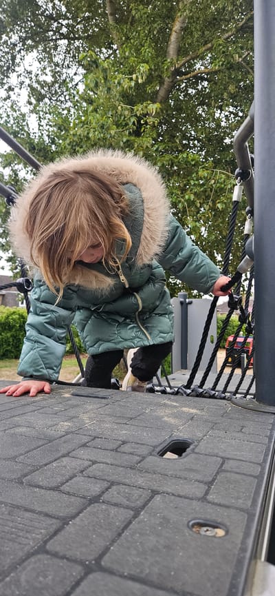 A child wearing a green quilted winter coat with a fur-trimmed hood is climbing on a black rope structure attached to a gray brick surface with square openings. The setting appears to be a playground or park with greenery and trees visible in the background. In the distance, a red vehicle and other park structures are discernible. The image captures a moment of the child's activity within the playground environment. The city of Norwich, United Kingdom is not discernible from the image.