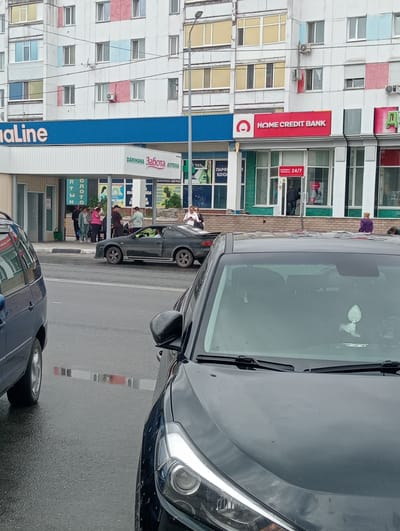 A street view captures a black sports car parked on the side of the road in front of a row of businesses in Kostanay, Kazakhstan. A dark blue minivan is parked to the left of the sports car, partially obscuring it. The facade of a building is visible behind the cars, featuring storefronts including "HOME CREDIT BANK," "DARİHANA," and "Забота АПТЕКА." Several people are standing on the sidewalk in front of the businesses, some interacting with each other. The street and sidewalk appear wet, suggesting recent rain. The overall scene is one of urban street life, with vehicles and pedestrians present.
