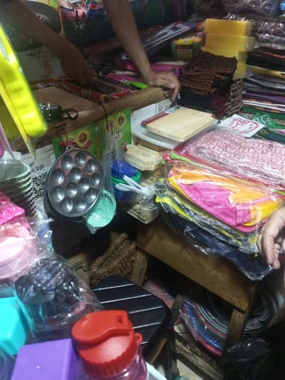 A cluttered vendor stall presents a variety of household items. Wooden cutting boards, colorful cloths wrapped in plastic, and a metal mold for making small cakes are prominent. Plastic containers and kitchen tools are also visible. Stacks of fabrics and yellow sponges fill the background, with a person's hand visible amidst the merchandise. The scene suggests a bustling market environment.