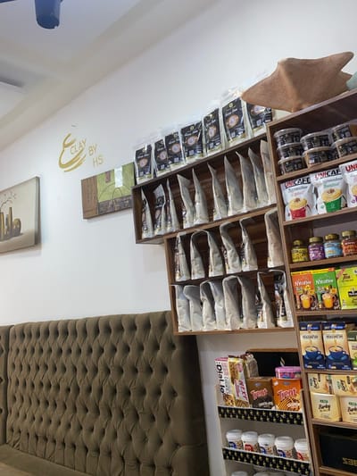 This image depicts the interior of a commercial establishment in Kabala, Nigeria. The scene is characterized by white walls adorned with minimalist decor and extensive product shelving.
On the left wall, "CLAY BY HS" is inscribed in stylized golden lettering, likely Arabic calligraphy. Below this, two framed artworks are visible: a muted painting of several vases on a table to the far left, and an abstract piece featuring earthy tones and geometric shapes. A long, plush, tufted upholstered bench or sofa, in an olive-green or dark brown hue, lines the lower portion of the left wall.
To the right, two interconnected wooden shelving units are packed with various consumer products. The upper and middle sections of the left shelving unit display numerous clear plastic pouches containing a light-colored granular product, identified by labels reading "Garri Kowa." The right, vertical shelving unit showcases a diverse array of packaged goods across multiple tiers:
* The top tier holds small, round, dark-colored containers (some labeled "UNICAFE"). A reddish-brown, abstract decorative object rests on the topmost shelf.
* Lower tiers display various coffee products, including "UNICAFE Espresso Macchiato," "NV Coffee" sachets, and "Marami 3in1 Coffee." Also present are "KADAK Tea" packages, jars of "HANDY PUMPKIN SPICE," and different brands of packaged cookies such as "Dialo Jo," "NEXT," and "Treat." The lowest visible shelves contain additional white plastic containers.
No individuals are present within the frame. The lighting appears to be provided by ceiling-mounted fixtures, one of which is partially visible at the top left.