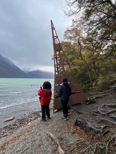 Two individuals stand on a metallic structure at the edge of a pebble beach beside a body of water. One person, clad in a red jacket, faces the water and appears to be holding a smartphone, possibly taking a picture of the mountain landscape visible in the distance. The other person, wearing a black jacket and beanie, stands closer to the viewer. An A-frame structure with a sign is situated on the left. Trees and vegetation are seen in the background. The sky is overcast, and the water seems a pale green color. It seems this scene is near the coast of the city, Lapataia, Argentina.