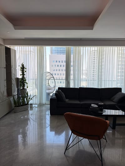 This image displays a bright, modern living space within a high-rise apartment or condo, characteristic of urban environments like Lusail, Qatar. The scene is dominated by a large window overlooking a city skyline, bathed in natural light, indicating it is daytime on a clear day.
A dark, contemporary sectional sofa is positioned in the foreground, facing the window. Adjacent to it, a distinct orange upholstered armchair with slender black metal legs provides a vibrant contrast. A small, dark side table next to the sofa holds a cup and a tissue dispenser.
The expansive window is draped with sheer white curtains that softly filter the sunlight, yet allow views of other tall buildings. Through the window, a spherical, possibly wicker, outdoor chair can be seen on a balcony. The floor is covered in polished light gray tiles, highly reflective, mirroring the curtains, furniture, and light from the windows.
Along the wall beneath the window, a white credenza or cabinet is visible. On its surface, two decorative planters house healthy green plants; one is a vine climbing a moss pole, and the other is a broad-leafed plant. The room's ceiling is light-colored and minimalist, contributing to the clean aesthetic.
No people are present, and no specific activity is taking place, suggesting a serene and unoccupied environment. The decor is modern and minimalist, emphasizing clean lines and natural light. No discernible text is visible on any objects or surfaces within the frame.