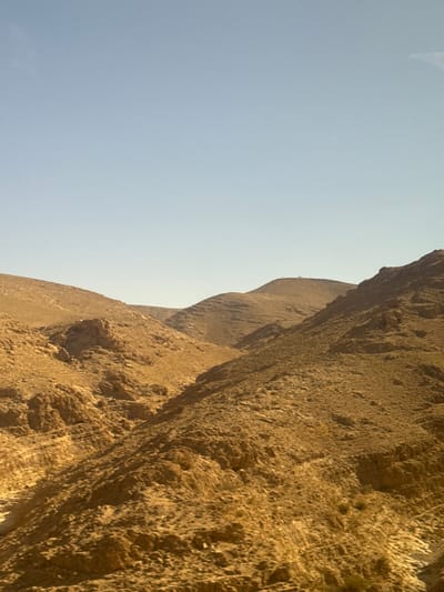 The image depicts a dramatic natural landscape dominated by a steep, rocky cliff face under a clear, pale blue sky. The cliff, rendered in shades of brown and tan, shows signs of erosion and has a rough, textured surface with visible striations and crevices. In the far distance, along the edge of the cliff, a line of small figures suggests human presence, possibly hikers or climbers, dwarfed by the immense scale of the natural formation. The lighting indicates it is daytime, likely morning or afternoon, with the sun casting subtle shadows that highlight the rugged topography. The overall scene evokes a sense of natural grandeur and isolation. The crops provide closer views of the rock texture and the vastness of the sky. No text is visible in the image.