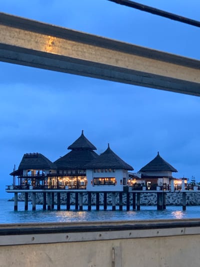 This image depicts a tranquil coastal scene in M'diq, Morocco, likely at dusk or early night. The primary subjects are several charming overwater bungalows or restaurants, each featuring distinctive, multi-tiered peaked roofs, possibly thatched, and white walls. These structures are built on sturdy piers or stilts that extend into a calm body of water, which reflects the ambient light.

The sky is a clear, deep blue, indicating the transition from twilight to night. Warm, inviting lights glow from inside the buildings, illuminating their interiors and creating a serene atmosphere. The architectural style suggests a resort or leisure setting. In the foreground, a large, vertical metallic beam or structure is partially visible on the left, hinting that the photo was taken from an adjacent platform, bridge, or boat. The water appears still, contributing to the overall sense of peace. No people or specific activities are discernible, and there is no visible text. The scene evokes a feeling of relaxation and quiet beauty.