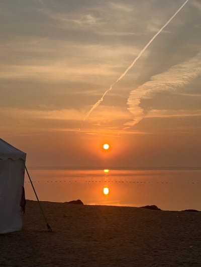 A sunrise casts a warm, orange glow over a tranquil body of water, reflecting the sun's brilliant orb. The scene is set on a sandy beach, with a white tent visible on the left, its green support pole anchored to the ground. In the distance, a faint line of buoys stretches across the water. The sky is a canvas of soft clouds, streaked with a long, white contrail from an aircraft. The overall impression is one of peaceful solitude in Surakovo, Russia.