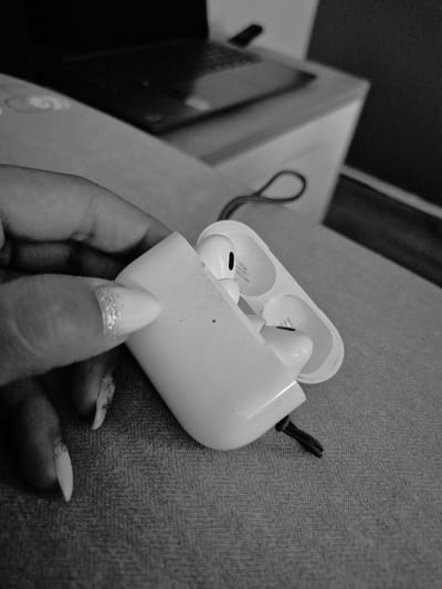 The image shows a close-up, black and white shot of a person's hand holding an open white earbud case. Two white earbuds are visible inside the case. The person's nails are long and decorated with glitter. The background shows a blurred surface, possibly a sofa or bed, and a hint of a laptop and furniture. There is no identifiable information in the image that would allow for determining the location in Iskrets, Bulgaria.