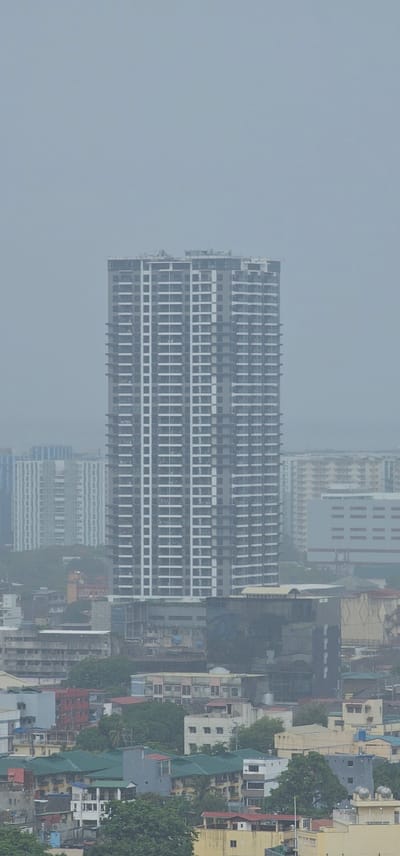 This media file is an aerial photograph of a dense urban landscape in Makati, Philippines, under hazy or overcast conditions, suggesting either early morning, late afternoon, or a day with poor visibility. The perspective is from a high vantage point, looking down at a city teeming with buildings of various sizes and architectural styles.

The most prominent subject is a large, modern, multi-story building with a uniform facade of gray and white. It features rows upon rows of windows, indicative of a residential or office complex. Surrounding this main structure is a more varied collection of buildings, including older, more traditional edifices with colorful roofs (some green, some red), and other contemporary high-rises. Trees are interspersed throughout the cityscape, adding patches of green against the predominantly gray and beige tones of the built environment.

The overall scene depicts a bustling city with signs of ongoing development, as evidenced by the construction of the large central building. There are no people visible, and the focus is purely on the urban sprawl. The weather appears to be contributing to the muted color palette and lack of sharp detail, with a diffused light. There is no text readily legible in the image, though a sign with "SOUTHEASTERN COLLEGE" is partially visible on one of the buildings in the upper right section of the image. The overall impression is one of a dynamic, working metropolis.