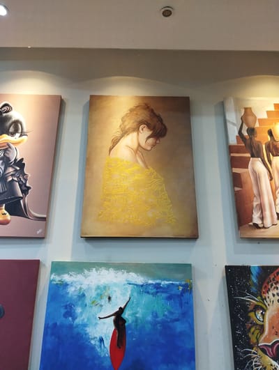 The image depicts a wall displaying several framed paintings. From left to right, the top row features a painting of a cartoon character, possibly Donald Duck, followed by a portrait of a woman with her back to the viewer, draped in a yellow garment. To the right of the woman's portrait is a painting of two figures carrying pottery on steps. In the bottom row, a painting with a predominantly blue and white abstract design, featuring a figure on a red surfboard, is visible. To its right is a close-up painting of a leopard's face. The paintings are mounted on a light-colored wall, and a ceiling light fixture is visible at the top of the image. The scene appears to be an indoor art display, potentially in a gallery or shop. The presence of an art gallery with such diverse artwork in Hurghada, Egypt, suggests a tourist-oriented location where local and international art may be showcased.