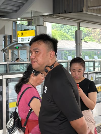 In the city of Longta, China, a man wearing a black t-shirt and a neckband is seen in the foreground. He is looking to his left, with a slight smile. Behind him, a woman in a pink shirt with a red sash around her waist is also looking to her left. To the right of the man, a woman in a black t-shirt is holding a red object, possibly a phone, and looking down. The scene appears to be at a transit station, with signs indicating "Exit" and "Toilet" with arrows pointing in different directions. The environment includes trees and buildings in the background, suggesting an outdoor or semi-outdoor setting. The lighting is bright, indicating daylight.