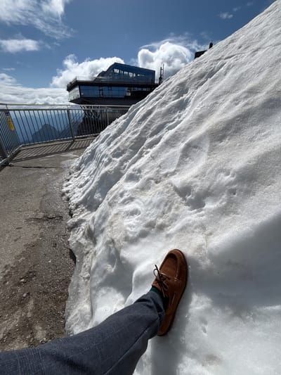 This is a first-person perspective, eye-level medium shot of a person's legs and feet near a large snowdrift at the Ehrwald, Austria. The person is wearing dark gray checkered trousers and a brown suede boat shoe with green socks. The snowdrift occupies the right two-thirds of the frame and shows signs of melting, with some patchy dirt and water visible at its base. To the left of the snowdrift, a paved walkway is visible, speckled with dirt and gravel, and wet in places. A metal railing runs along the edge of the walkway, separating it from a steep drop-off. In the background, a modern building with large glass windows, a restaurant or observation deck, is perched on the mountainside. The sky above is blue with scattered white clouds. The scene appears to be a scenic mountain location.