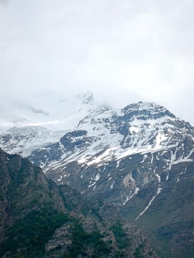 This is a vertical, medium shot of a snow-capped mountain under a cloudy sky. The mountain has a rocky, dark gray face with patches of green vegetation on the lower slopes. Snow covers the upper portions of the mountain, with some rocky outcrops visible through the snow. A glacier is visible on the left side of the mountain. The sky is overcast and white. The image does not depict the city of Gemur, India.