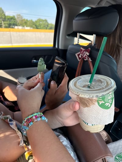 The image captures an interior view of a vehicle, likely in motion, with a focus on the passengers and their immediate surroundings. The scene is lit by daylight filtering through the car windows. A Starbucks iced beverage with a green straw and sleeve is held by a hand in the upper right quadrant, alongside a brown leather purse. Another person is holding a phone and a green animal-shaped lollipop. The person holding the lollipop also has multiple colorful bracelets on their wrist. The background reveals a road and trees outside the car window. There is no way to discern the location to be Santa María Moyotzingo, Mexico.