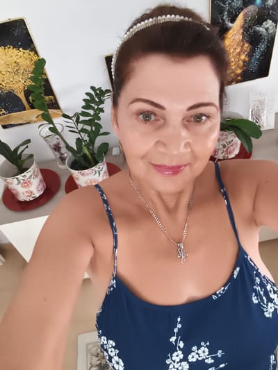 An adult Caucasian female with brown hair and green eyes, wearing a pearl headband, a silver necklace with a cross pendant, and a dark blue floral print dress, is taking a selfie in what appears to be a domestic interior. The background features several framed pieces of art on the wall, one depicting a golden tree against a starry night, and another with a golden-orange crystalline texture. Two potted plants with floral designs on their pots are visible on a surface in the middle ground. The lighting suggests an indoor setting, possibly a living room or dining area. There is no specific indication of the location being in Livorno, Italy.