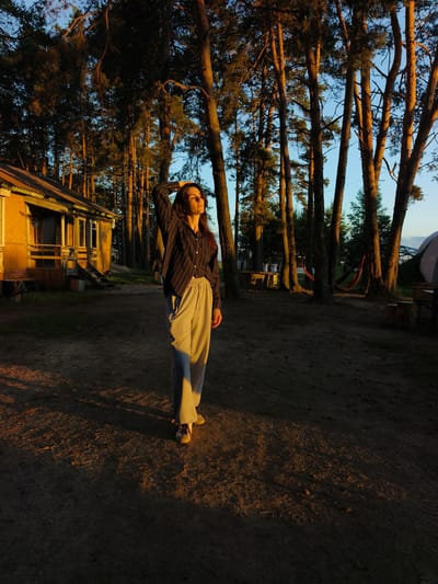 A young woman stands in a dirt clearing next to a yellow wooden building with a porch, surrounded by tall pine trees. The sun is setting, casting long shadows and bathing the scene in warm golden light. The woman is wearing a striped shirt and gray sweatpants, with one hand raised to her head. In the background, there are picnic tables, a hammock, and a tent. The overall impression is of a serene, rural campsite in Онохино, Russia.