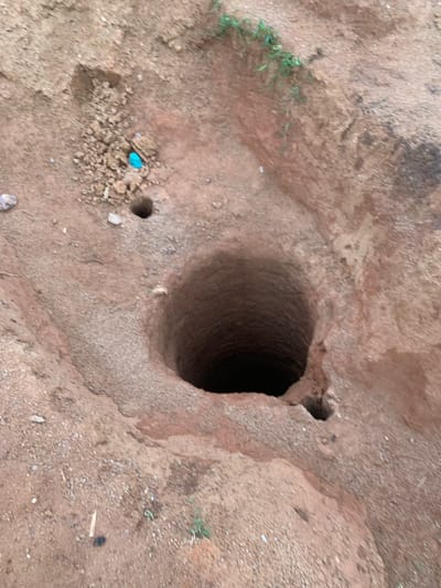 In Buga da Gindi, Nigeria, a circular well, approximately one meter in diameter, has been excavated in reddish-brown soil. The well descends into darkness, with its depth not discernible from the image. Surrounding the well are the excavated soil mounds, creating an uneven terrain. A small pile of loose dirt is located to the left of the well, and a bright blue object, possibly a piece of plastic, rests atop it. A smaller, darker hole, approximately 10 centimeters in diameter, is visible near the rim of the main well. Sparse green vegetation grows along the upper edge of the excavated area.