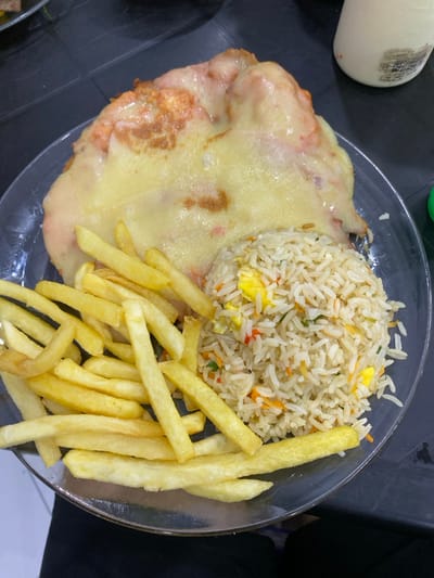 A plate of food sits on a dark surface. It features a portion of fried rice molded into a dome shape, alongside a serving of golden French fries. A piece of meat is covered in melted cheese. A white sauce container is visible in the background. The plate appears to be clear glass or plastic.