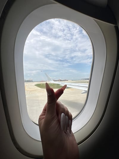 A close-up view from inside an airplane shows a person's hand forming a heart shape with their thumb and index finger against the window. Outside the window, an airport tarmac is visible with an airplane wing and tail in the background. The sky is partly cloudy. The image does not depict any recognizable landmarks or features that would allow for identification of a specific location within Ta Khmau, Cambodia.