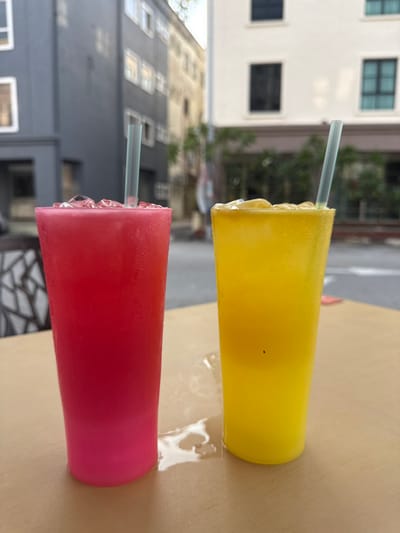 Two tall, tapered plastic cups containing iced beverages are positioned on a light brown table in Malacca City, Malaysia. The left cup holds a magenta-red drink, and the right cup holds a bright yellow drink, both topped with ice and containing clear plastic straws. Condensation is evident on the exterior of both cups, with liquid pooling around their bases and between them. A small dark speck is visible on the side of the yellow cup. The blurred background depicts an urban street scene with multi-story buildings; a dark grey structure is visible to the left, and a cream-colored building with dark windows is to the right. Green foliage is indistinctly visible between the structures. The scene is illuminated by ambient daylight. No individuals are visible.