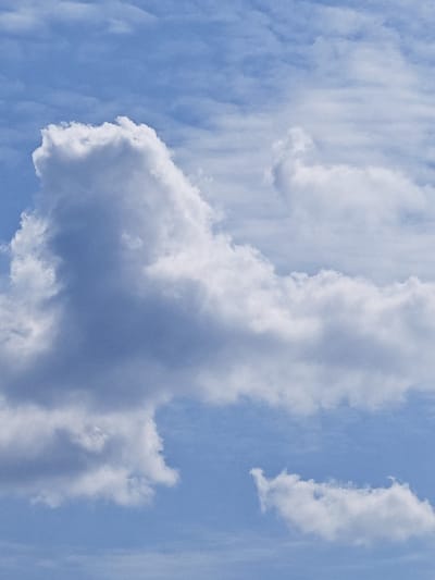 A bright blue sky is filled with fluffy white clouds. The clouds are scattered across the sky, with some appearing larger and more defined than others. One large cloud formation in the center of the image is particularly prominent, resembling a vaguely animalistic shape. In the lower right of the image, another smaller cloud formation is visible. The overall scene depicts a clear and pleasant day. The provided location of Kurumoch, Russia, suggests this is an outdoor scene, possibly a landscape or skyscape captured in that region.